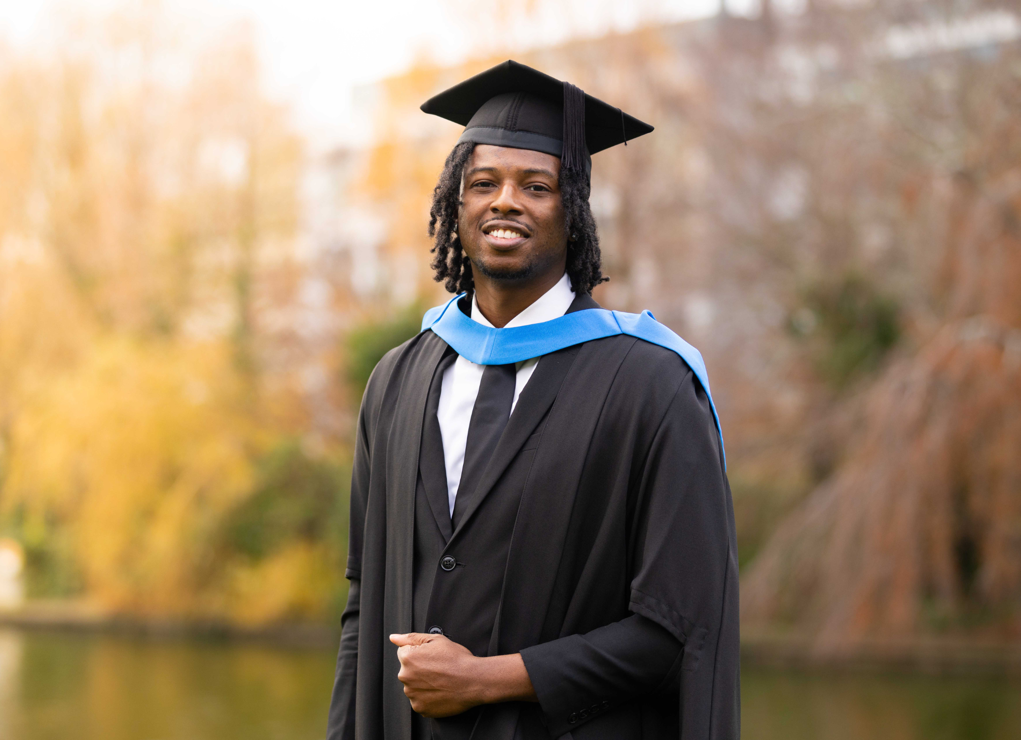 UCD alumnus Segun Ologundudu wearing graduation cap and gown, posing for a photo by the main UCD lake.